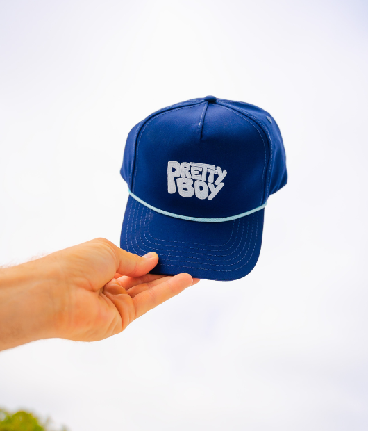 Hand holding blue hat with "Pretty Boy" in white letters and white rope on brim