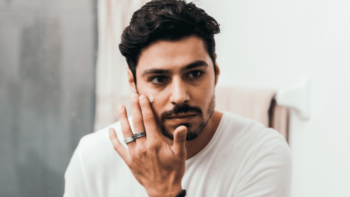 Man inspecting skin in a bathroom mirror, applying moisturizer to his cheek