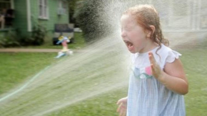Child reacting to water spray from a lawn sprinkler in a grassy yard.