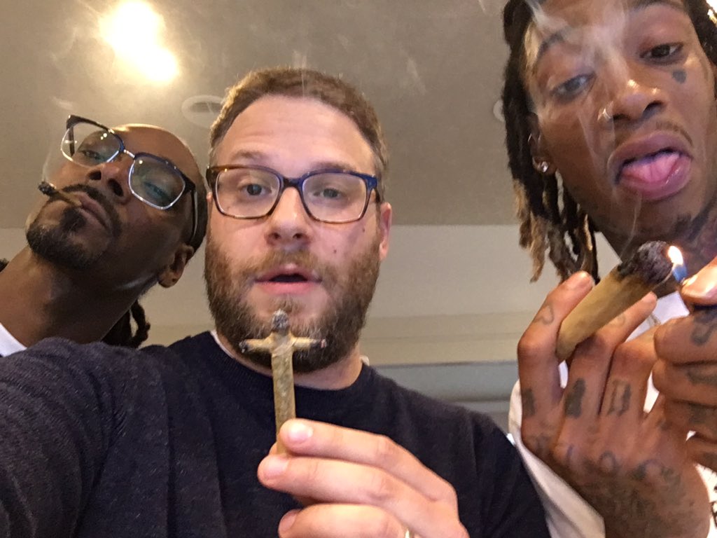 Three men, one holding a cross-shaped joint, another lighting a large joint, smoke visible around them