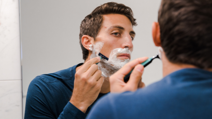 Man shaving his face with a razor and shaving cream in front of a bathroom mirror