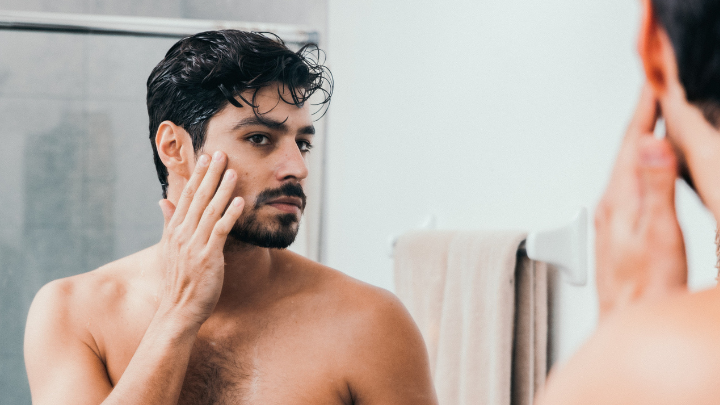 Man examining his face in a bathroom mirror, possibly checking for acne or skin issues.