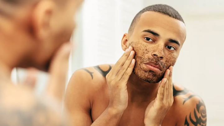 Man applying facial scrub to exfoliate skin, looking in mirror
