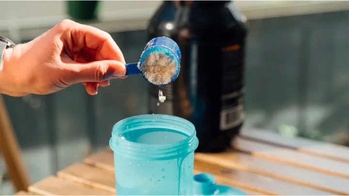 Hand holding a blue scoop of powder above a blue shaker bottle on a wooden table