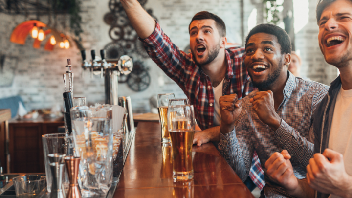 Three men at a bar cheering excitedly with glasses of beer in front of them