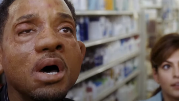 Man with severely swollen face and eye, standing in a store aisle with a woman in the background