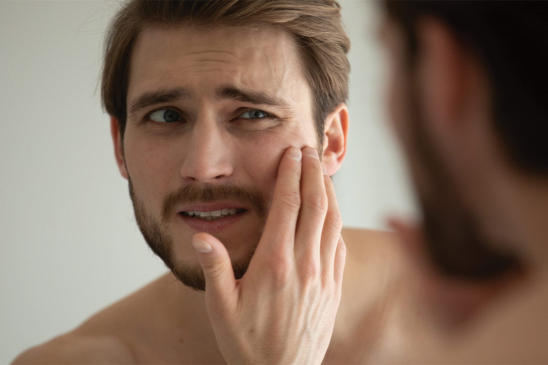 Man with beard touching his cheek, looking concerned while examining his face in a mirror