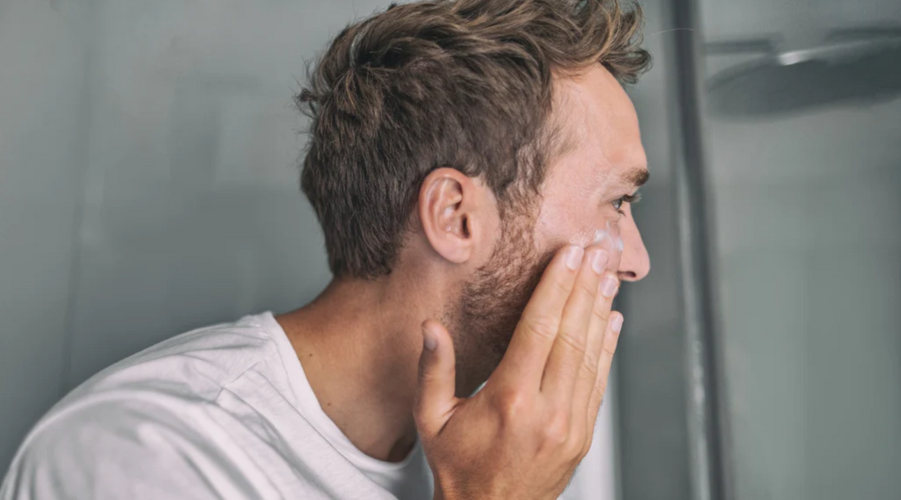 Man applying cream to his face in a bathroom mirror, illustrating skincare routine for rosacea