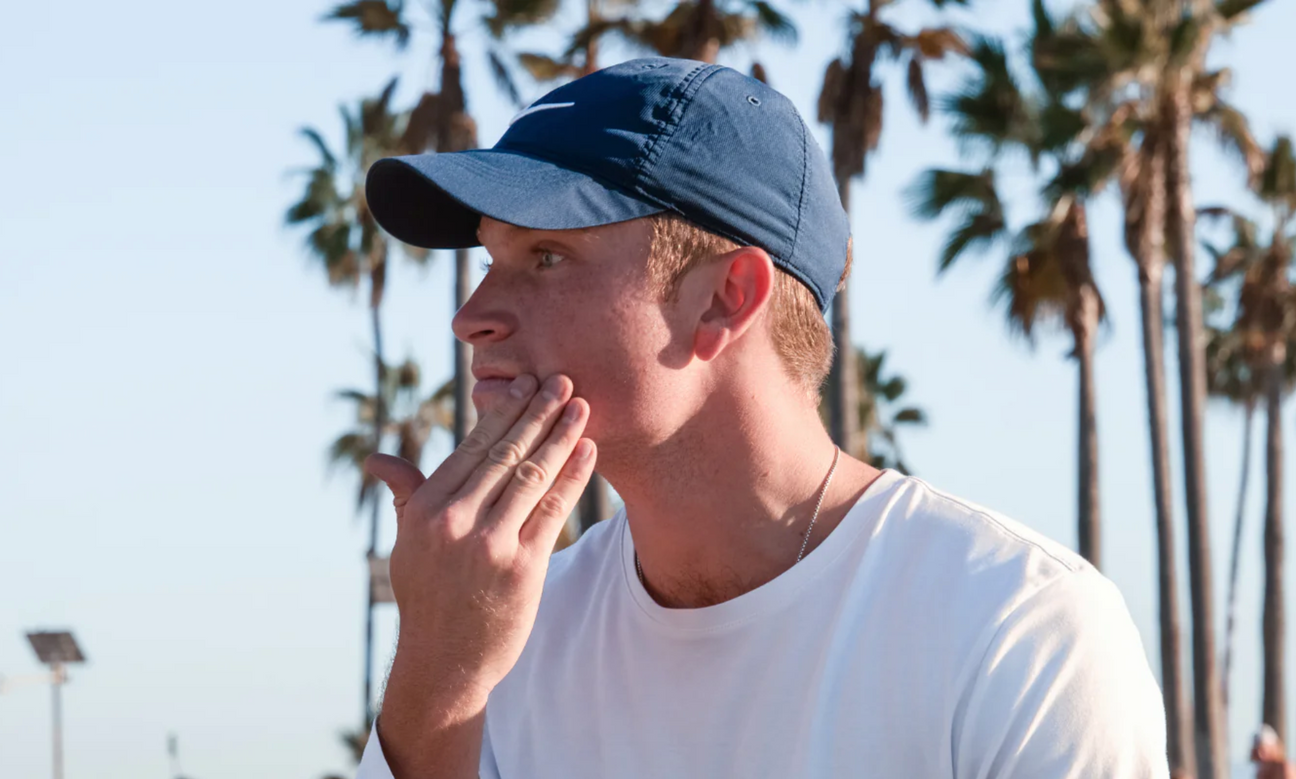 Man in a blue cap touching his cheek outdoors with palm trees in the background