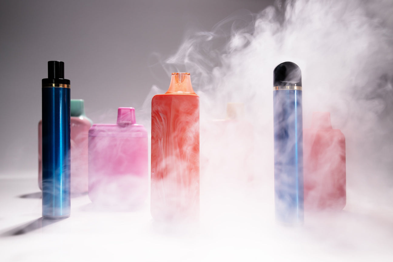 Various colorful vape devices surrounded by vapor smoke on a white surface