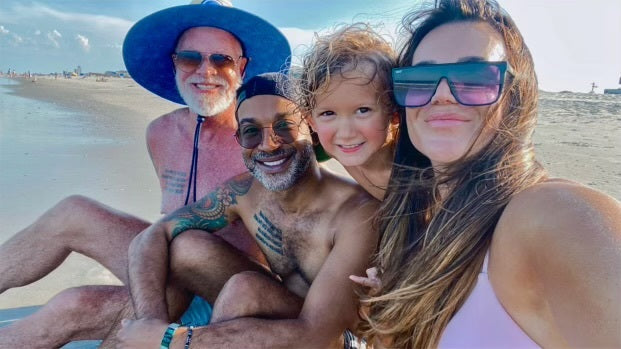 Four people smiling at the beach, including an older man with a blue hat, a man with tattoos, a young child, and a woman with large sunglasses